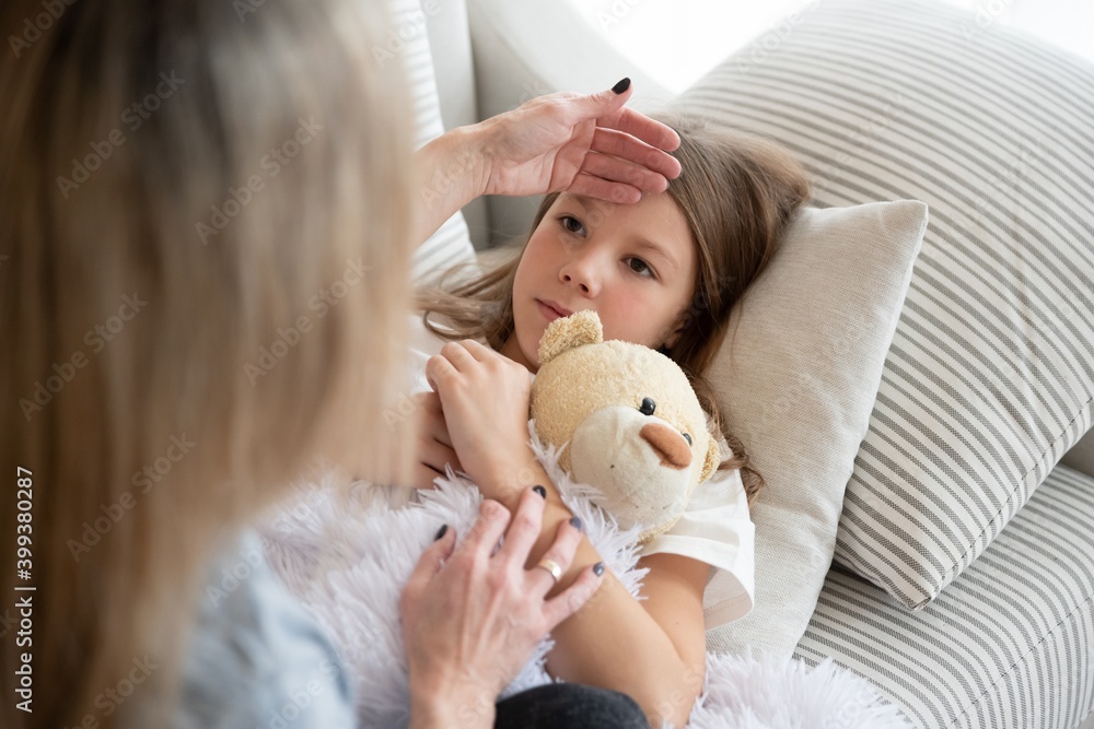 The sick girl is lying with the teddy bear. Mom is checking for a fever.