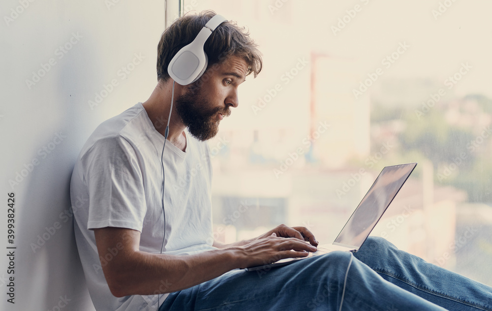 bearded man with an open laptop sits on the windowsill and headphones on his head bearded man with an open laptop sits on the windowsill and headphones on his head