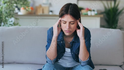 Video of frustrated young woman with headache and frowning while sitting on couch in the living room at home.