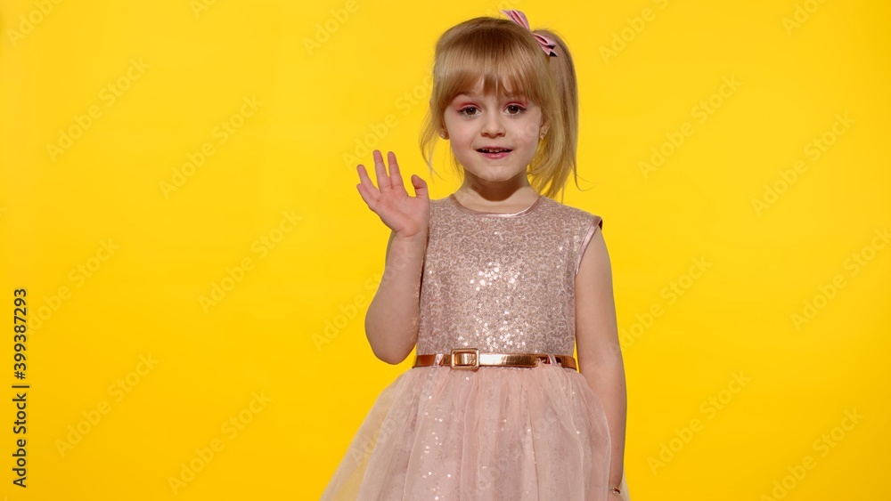 Friendly little blonde child kid girl 5-6 years old with fashion makeup in pink shiny dress, waving to camera doing hello or bye gesture isolated on yellow background. Friendly little blonde child kid girl 5-6 years old with fashion makeup in pink shiny dress, waving to camera doing hello or bye gesture isolated on yellow background.