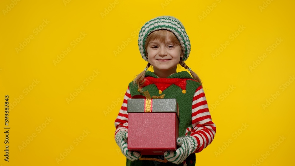 Little teen kid girl in Christmas elf Santa helper costume isolated on yellow background. Child giving present gift box to camera, smiling sweetly. People New Year holidays celebration Little teen kid girl in Christmas elf Santa helper costume isolated on yellow background. Child giving present gift box to camera, smiling sweetly. People New Year holidays celebration