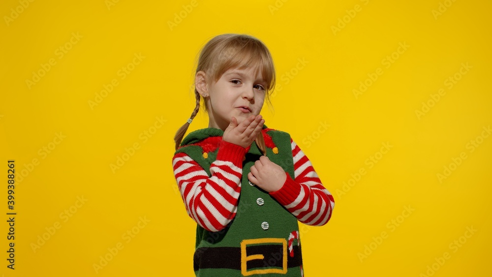 Naklejka premium Shy shamed child girl in Christmas elf Santa helper costume posing looking camera blows air kiss. Little kid feels guilty. Isolated on yellow background studio. People emotions. New Year holidays
