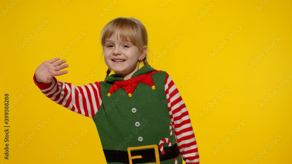 Fototapeta premium Teen kid girl 5-6 years old in Christmas elf Santa helper costume waving greeting with hand as notices someone. Hello, hi, greetings gesture. Small child laughing. People New Year holidays celebration