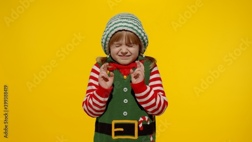 Shy smiling child. Christmas wish concept. Little fun blonde kid teen teenager girl in Christmas elf Santa helper costume hopes for dreams come, making a wish, fingers crossed, prays for better life