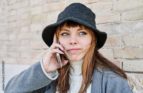 Portrait young pretty Caucasian girl, dressed in a gray jacket, a white sweater and a hat, talking on the mobile phone