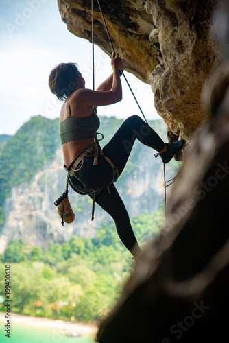 Confidence Asian woman climber with safety rope climbing high up on rocky mountain at tropical island in summer. Healthy and strong female enjoy active lifestyle and extreme sport in holiday vacation.