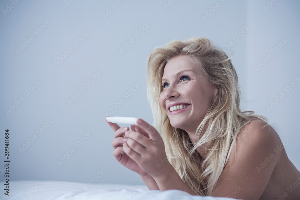 Smiling blond woman holding pregnancy kit while lying on bed at home Smiling blond woman holding pregnancy kit while lying on bed at home