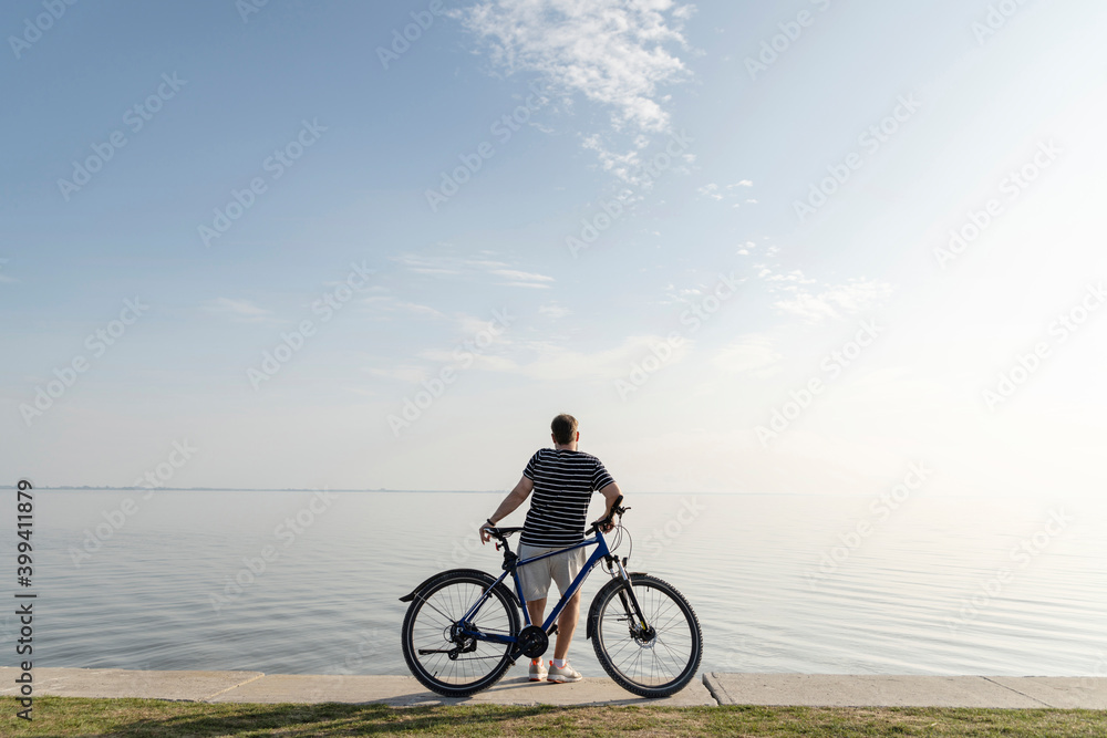 Man standing with bicycle looking at sea against blue sky Man standing with bicycle looking at sea against blue sky