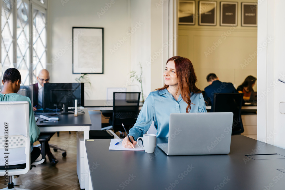 Smiling businesswoman writing while looking away in office Smiling businesswoman writing while looking away in office
