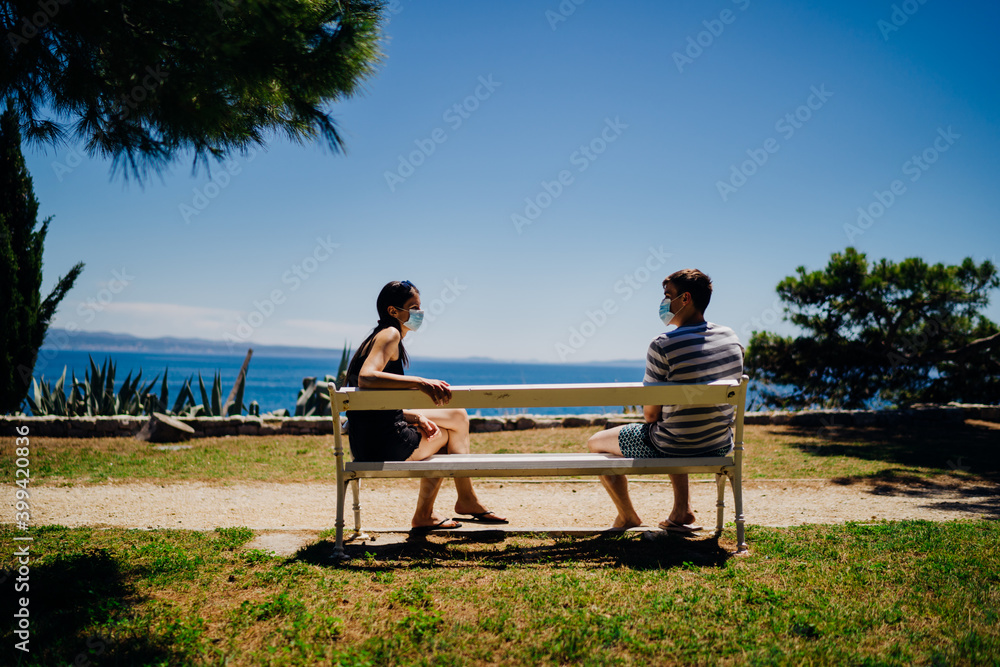 Young couple having an outdoor date on a bench in a park.Social distancing dating.Friends talking, wearing protective face masks.Coronavirus pandemic date.Meeting new people amid epidemic of COVID-19