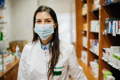 Friendly pharmacist working in a pharmacy amid coronavirus pandemic.Pharmaceutical health care professional providing COVID-19 therapy support.MPharm ina a drugstore wearing a protective face mask