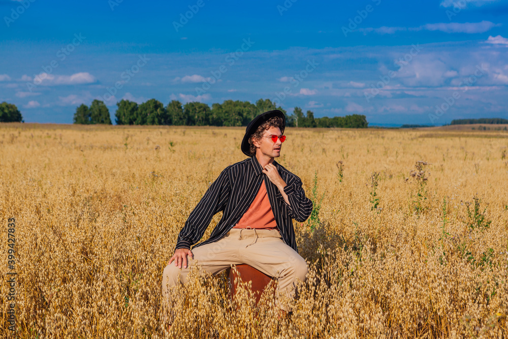 Tall handsome man with black hat and pink sunglasses sitting on a brown vintage leather suitcase at golden oat field. Tall handsome man with black hat and pink sunglasses sitting on a brown vintage leather suitcase at golden oat field.