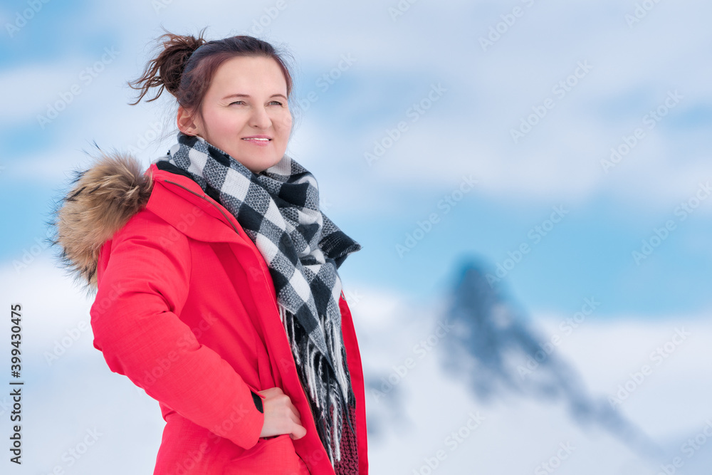 Portrait of woman traveler dressed in red winter windproof jacket, black and white scarf around neck. Mysterious lovely young woman on background of mountains and blue sky with white clouds.