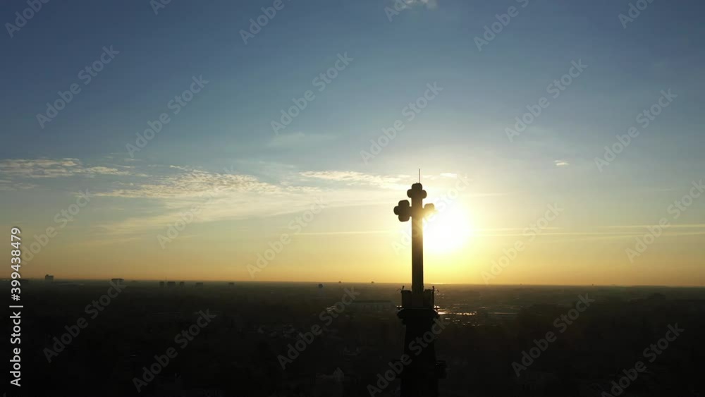 An aerial shot of a cathedral's steeple with a cross on top, taken at sunrise. The drone camera truck left around the cross. The golden sun shines behind it creating a silhouette.