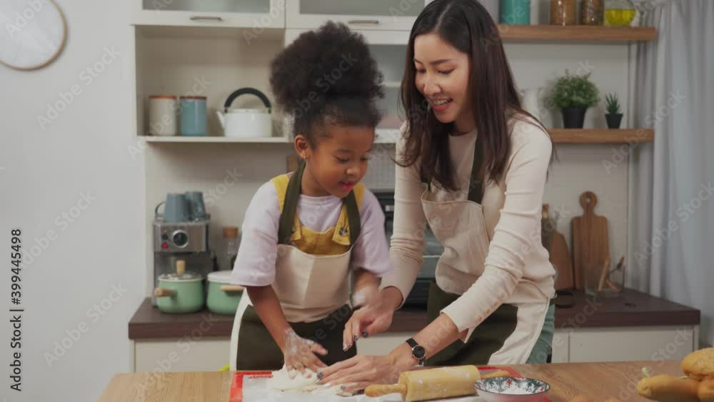 Happy beautiful Asian mother in apron standing at the kitchen table teaching little African daughter kneading a dough by hands, daughter know some technique by sprinkle the dough with a little flour Happy beautiful Asian mother in apron standing at the kitchen table teaching little African daughter kneading a dough by hands, daughter know some technique by sprinkle the dough with a little flour