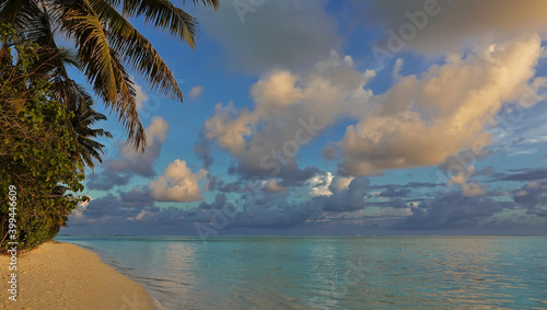 Fototapeta Naklejka Na Ścianę i Meble -  Golden hour in the Maldives. There are beautiful white and blue clouds in the azure sky. The aquammarine ocean is calm. The sandy beach has tropical vegetation.