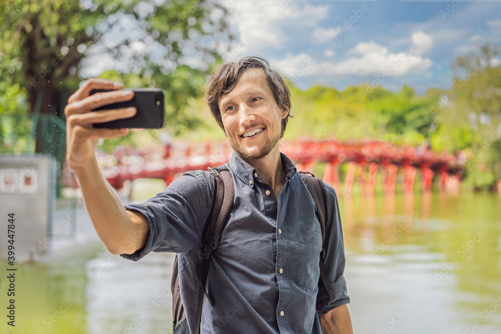 Fototapeta premium Caucasian man traveler on background of Red Bridge in public park garden with trees and reflection in the middle of Hoan Kiem Lake in Downtown Hanoi. Vietnam reopens after coronavirus quarantine COVID
