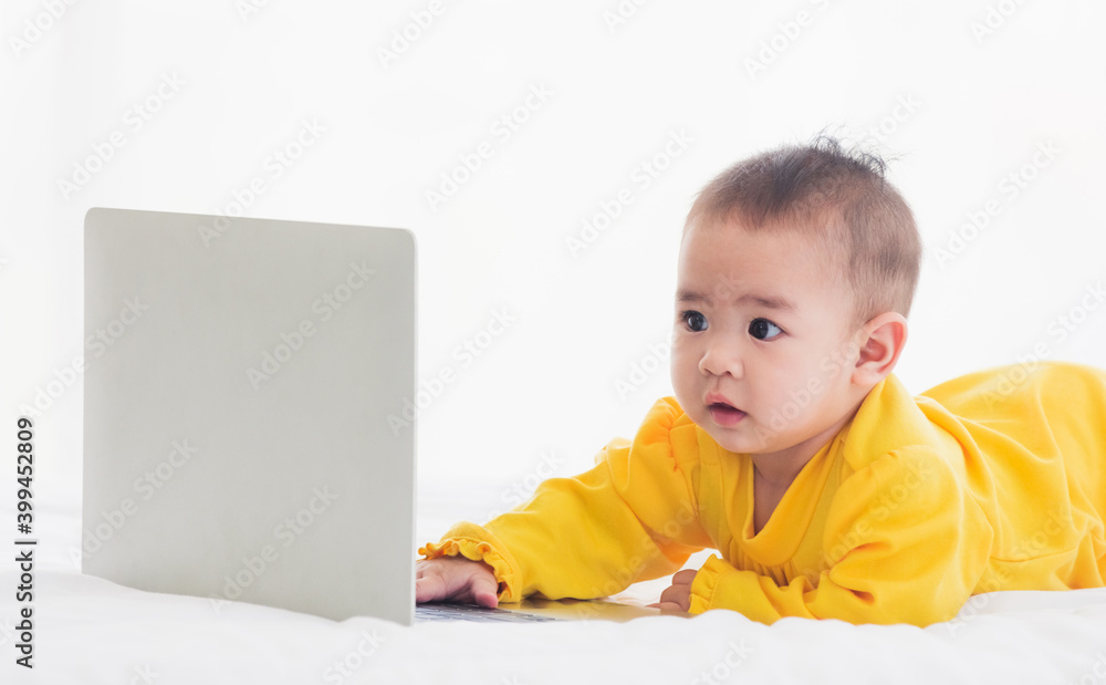 Asian little baby girl wearing a yellow dress typing hands on laptop computer keyboard and looking at monitor on the white bed. Little IT guy Asian little baby girl wearing a yellow dress typing hands on laptop computer keyboard and looking at monitor on the white bed. Little IT guy
