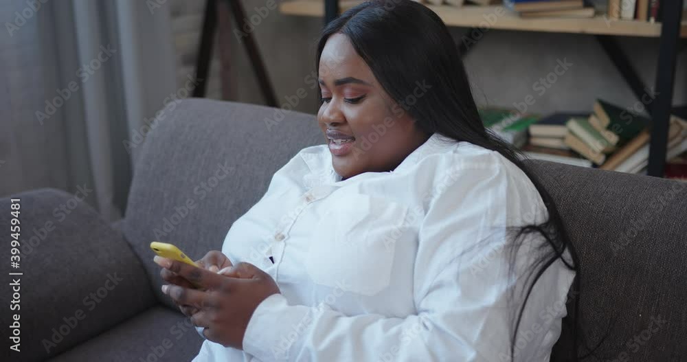 Overweight young African-American lady holds yellow smartphone and chats smiling and sitting on modern grey sofa close view Overweight young African-American lady holds yellow smartphone and chats smiling and sitting on modern grey sofa close view