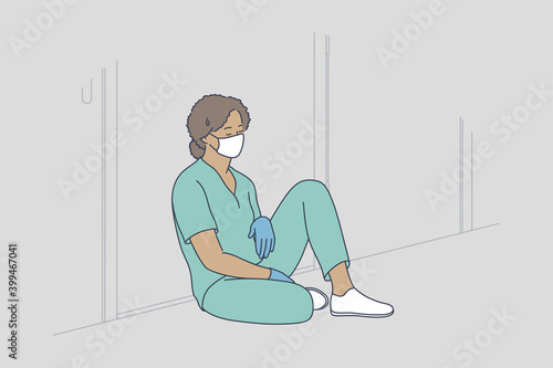 Doctor burnout, stress, hard conditions during pandemic concept. Tired exhausted black female nurse or doctor wearing protective face mask, gloves and blue uniform sitting on hospital floor