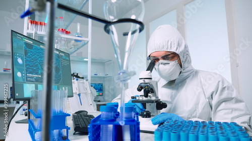 Laboratory scientist in ppe suit conducting experiment looking through a microscope in modern lab. Man working with various bacteria tissue, blood samples, concept of medical research for antibiotics