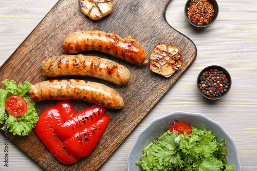 Wooden board with delicious grilled sausages, vegetables and spices on table