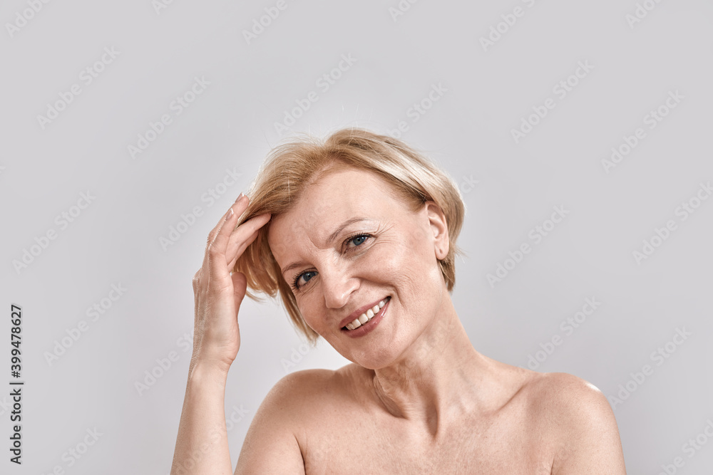 Fototapeta premium Portrait of beautiful middle aged woman smiling at camera, adjusting her hair, posing isolated against grey background. Beauty, skincare concept
