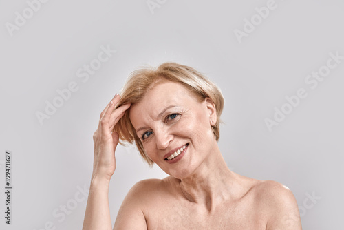 Portrait of beautiful middle aged woman smiling at camera, adjusting her hair, posing isolated against grey background. Beauty, skincare concept