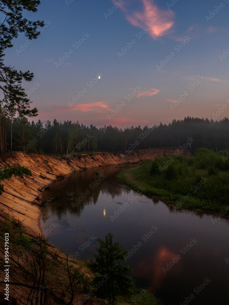 The clear evening sky and the Moon is reflected in the mirror surface of the forest river at sunset. Silence at sunset on the high bank of the forest river. The clear evening sky and the Moon is reflected in the mirror surface of the forest river at sunset. Silence at sunset on the high bank of the forest river.