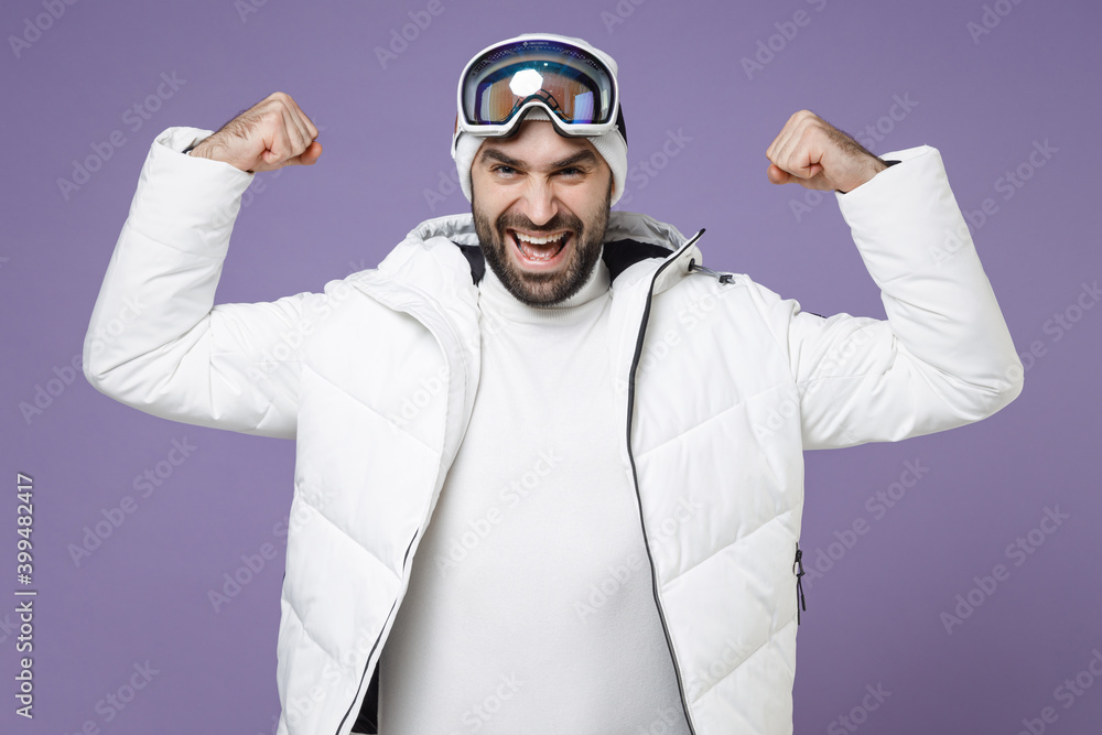 Strong skier man in white windbreaker jacket ski goggles mask showing biceps muscles spend extreme weekend winter season in mountains isolated on purple background. People lifestyle hobby concept. Strong skier man in white windbreaker jacket ski goggles mask showing biceps muscles spend extreme weekend winter season in mountains isolated on purple background. People lifestyle hobby concept.