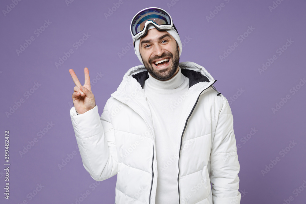 Naklejka premium Laughing skier man in white windbreaker jacket ski goggles mask showing victory sign spend extreme weekend winter season in mountains isolated on purple background. People lifestyle hobby concept.