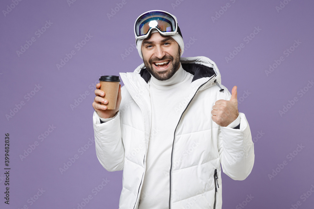 Funny skier man in white windbreaker jacket ski goggles mask hold paper cup coffee tea showing thumb up spend weekend winter in mountains isolated on purple background. People lifestyle hobby concept.