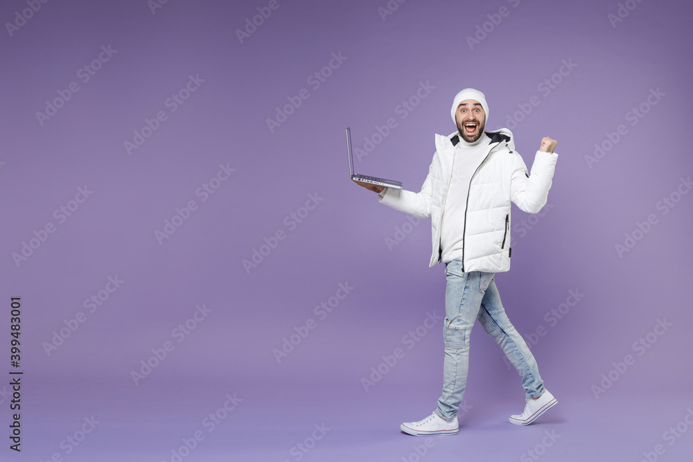 Full length side view of excited man in warm white padded windbreaker jacket hat working on laptop pc computer doing winner gesture isolated on purple background. People lifestyle cold season concept.