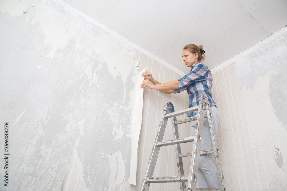 Young adult woman standing on metal ladder and tearing off old light wallpaper from wall in room. Making interior change. Preparing for home repair work. View from below.