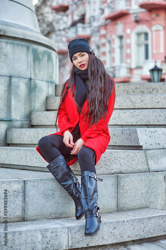 Beautiful young woman of Asian appearance on a winter city street . A girl in a red coat and a knitted hat