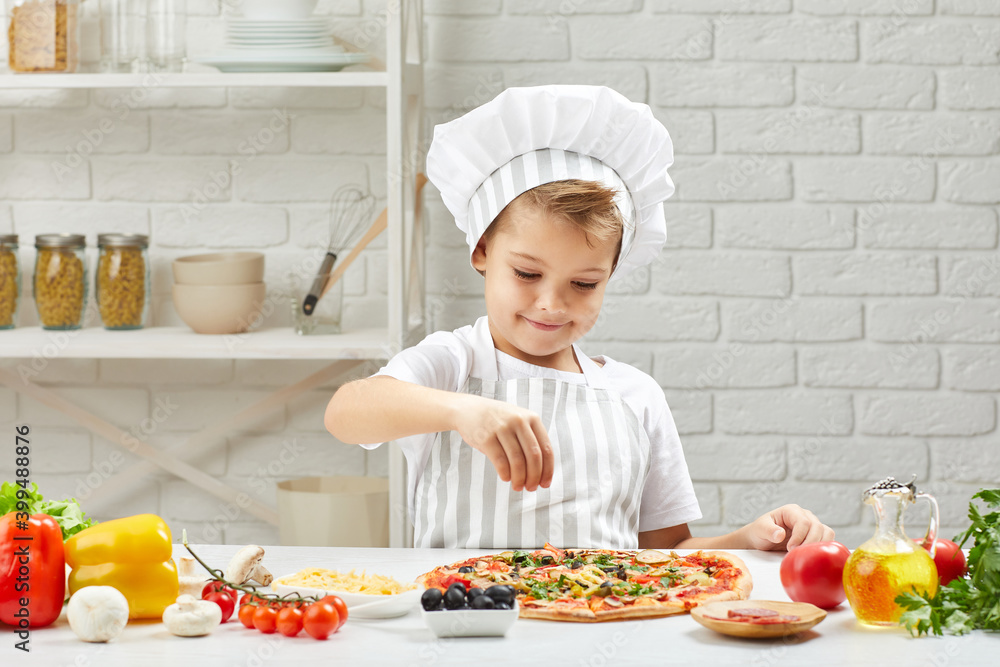 little boy in chef hat and an apron cooking pizza in the kitchen. child adding cheese into pizza little boy in chef hat and an apron cooking pizza in the kitchen. child adding cheese into pizza