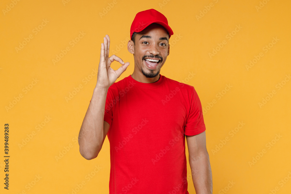 Delivery employee african man in red cap blank print t-shirt uniform workwear work courier service on quarantine covid-19 virus concept isolated on yellow background studio. Tattoo translation life. Delivery employee african man in red cap blank print t-shirt uniform workwear work courier service on quarantine covid-19 virus concept isolated on yellow background studio. Tattoo translation life.