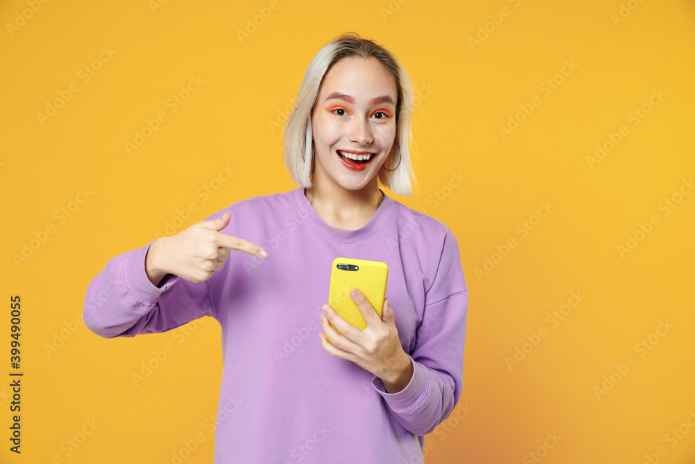 Young blonde smiling caucasian gladden woman 20s bob haircut wearing casual basic purple shirt pointing finger on mobile cell phone holding device isolated on yellow color background studio portrait Young blonde smiling caucasian gladden woman 20s bob haircut wearing casual basic purple shirt pointing finger on mobile cell phone holding device isolated on yellow color background studio portrait