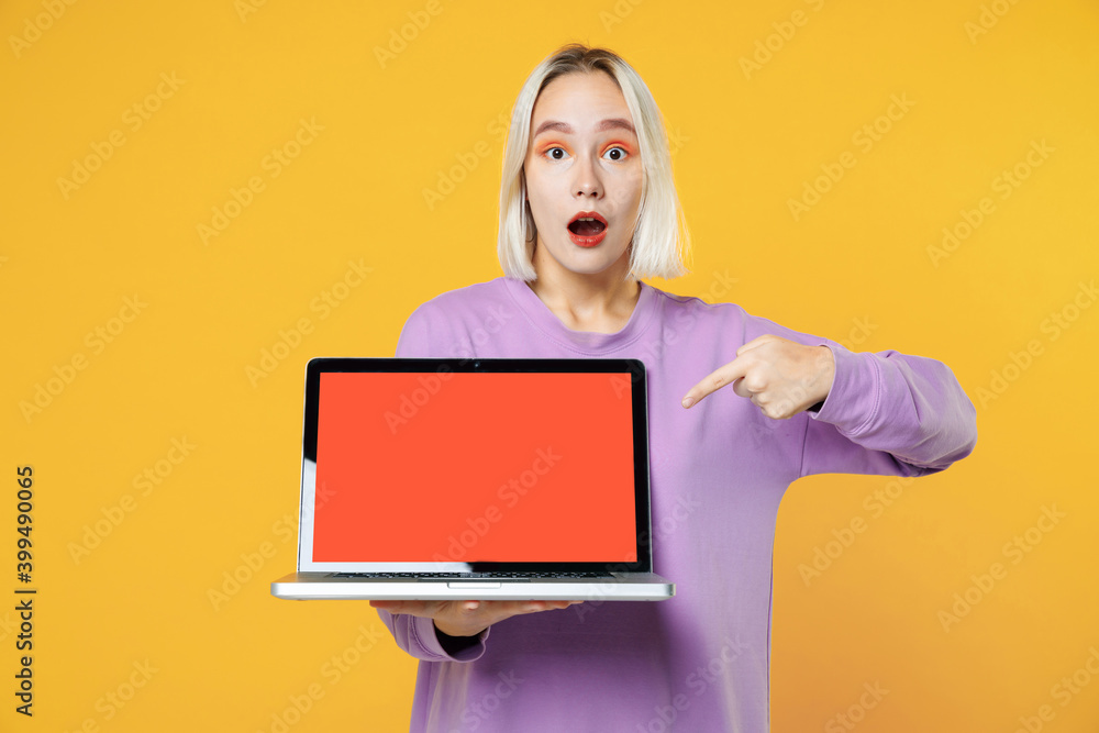 Shocked young blonde caucasian woman 20s in basic purple shirt using pointing finger on laptop pc computer with blank screen workspace copy space mock up isolated on yellow background studio portrait. Shocked young blonde caucasian woman 20s in basic purple shirt using pointing finger on laptop pc computer with blank screen workspace copy space mock up isolated on yellow background studio portrait.