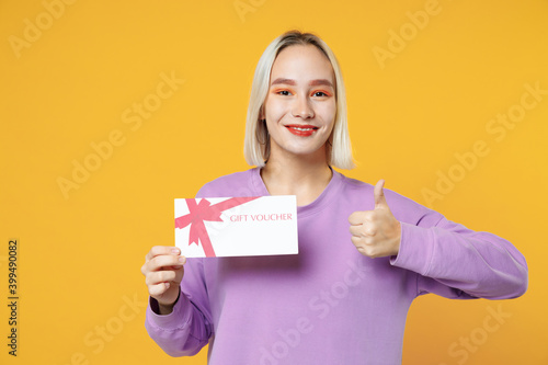 Excited young blonde caucasian woman 20s bob haircut bright makeup wearing casual basic purple shirt hold gift voucher mockup showing thumbs up gesture isolated on yellow background studio portrait.