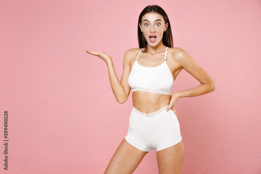 Shocked amazed attractive young brunette woman 20s in white underwear demonstrating sports fit body standing posing pointing hand aside isolated on pastel pink colour wall background, studio portrait. Shocked amazed attractive young brunette woman 20s in white underwear demonstrating sports fit body standing posing pointing hand aside isolated on pastel pink colour wall background, studio portrait.