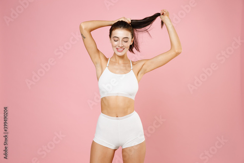 Smiling cheerful funny young brunette woman 20s in white underwear with sports body standing posing put hands on head making ponytail isolated on pastel pink colour background, studio portrait.