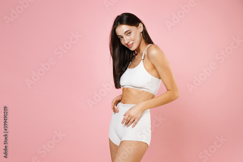 Side view of smiling beautiful young brunette woman 20s in white underwear with fit body posing looking camera hold hands on stomach belly isolated on pastel pink colour background, studio portrait.
