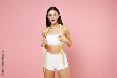 Attractive beautiful sports young brunette woman 20s in white underwear with sexy fit body posing hold measure tape looking camera isolated on pastel pink colour wall background, studio portrait.