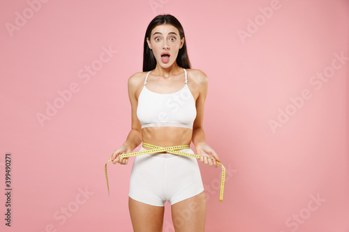 Shocked amazed beautiful sports young brunette woman 20s in white underwear posing measuring waist with measure tape keeping mouth open isolated on pastel pink colour wall background, studio portrait. © ViDi Studio
