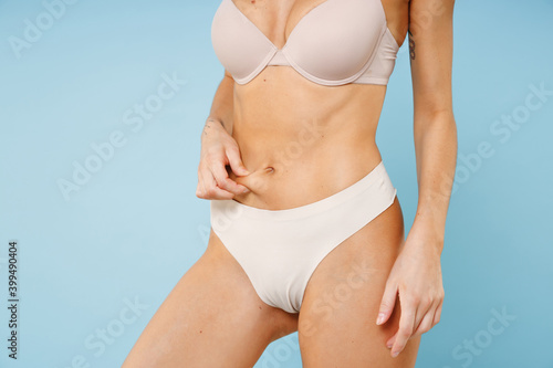 Cropped image of young brunette woman 20s in beige brassiere underwear showing sports fit body standing posing checking fat on her belly isolated on pastel blue colour background, studio portrait.