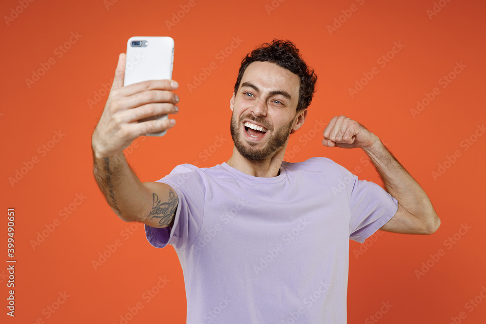 Cheerful young bearded man 20s wearing casual basic violet t-shirt standing doing selfie shot on mobile phone showing biceps muscles isolated on bright orange color wall background studio portrait. Cheerful young bearded man 20s wearing casual basic violet t-shirt standing doing selfie shot on mobile phone showing biceps muscles isolated on bright orange color wall background studio portrait.