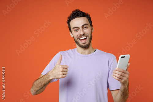 Laughing young bearded man 20s in basic violet t-shirt standing using mobile cell phone typing sms message showing thumb up looking camera isolated on bright orange color background studio portrait.