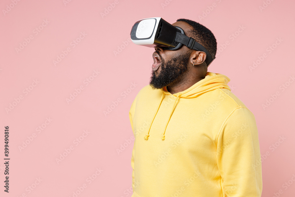 Shocked young african american man 20s in casual yellow streetwear hoodie watching in vr headset gadget rising keeping mouth open looking aside isolated on pastel pink wall background studio portrait. Shocked young african american man 20s in casual yellow streetwear hoodie watching in vr headset gadget rising keeping mouth open looking aside isolated on pastel pink wall background studio portrait.