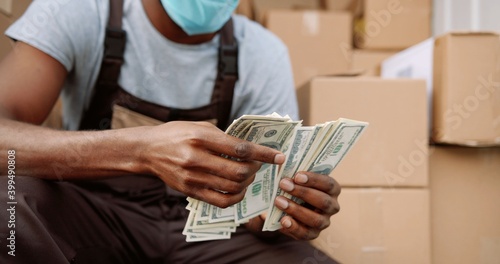 Close up of African American young handsome delivery man courier sitting in van with many boxes counting money dollars. Male postman holding money payment for shipment. Business concept